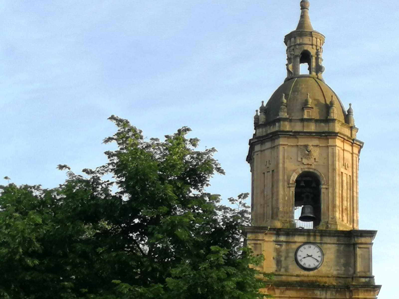 Sobre un cielo azul vibrante, el torreón de piedra de una iglesia barroca. Sobre la planta cuadrada pende un campanario octogonal abierto en el centro en un arco desde donde se ve la campana negra con un ribete dorado en lo bajo. Cierra la torre una cúpula decorada con varias formas cuadradas en la base y una pequeña torre coronada con una aguja. Frente a la iglesia, tapa la mitad de la imagen la copa de un gran árbol.