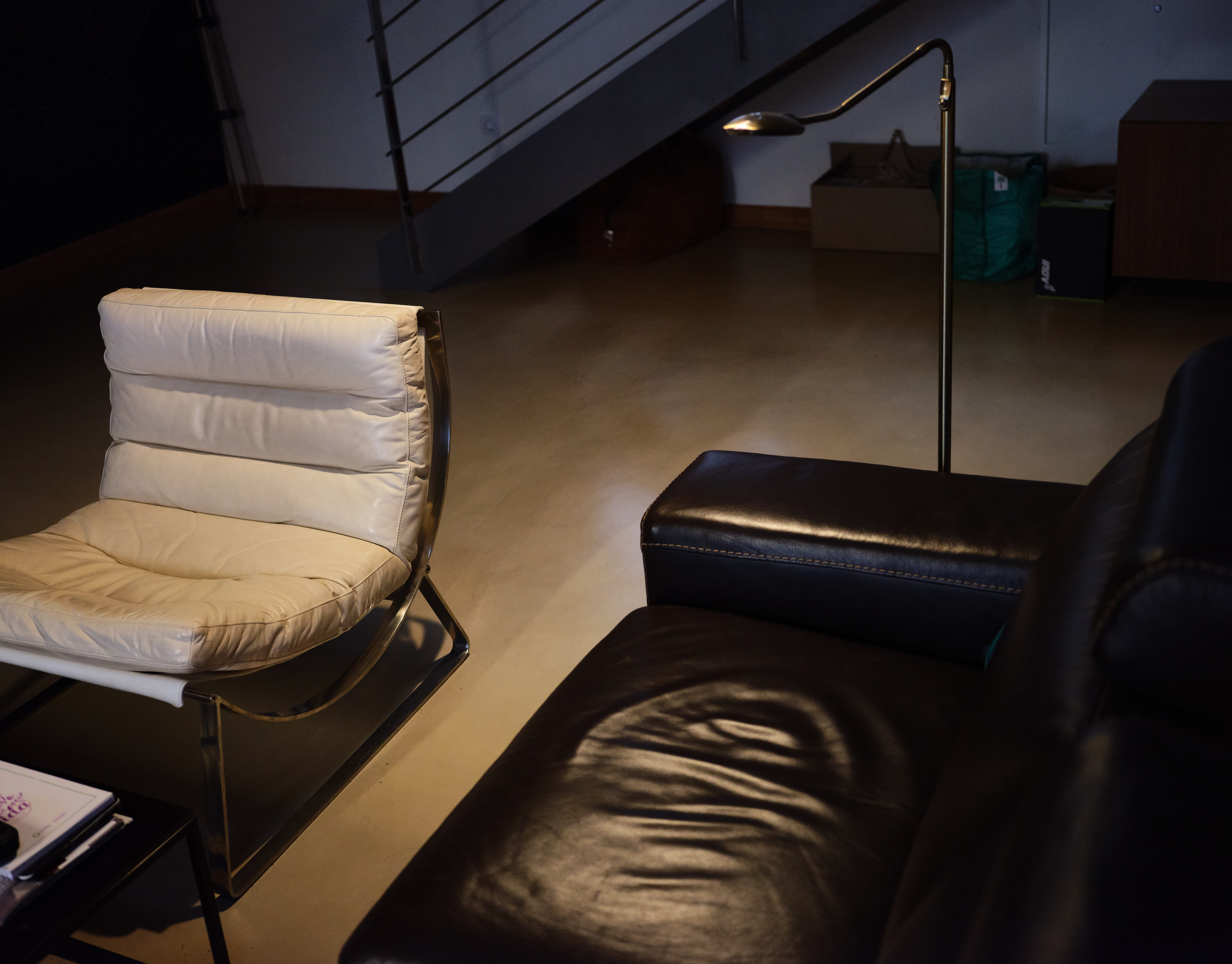 En un sótano iluminado por una lámpara se distinguen un sofá de cuero negro y una silla de metal cubierta con almohadillas blancas. Al fondo varias cajas y mochilas apiladas bajo unas escaleras de metal.